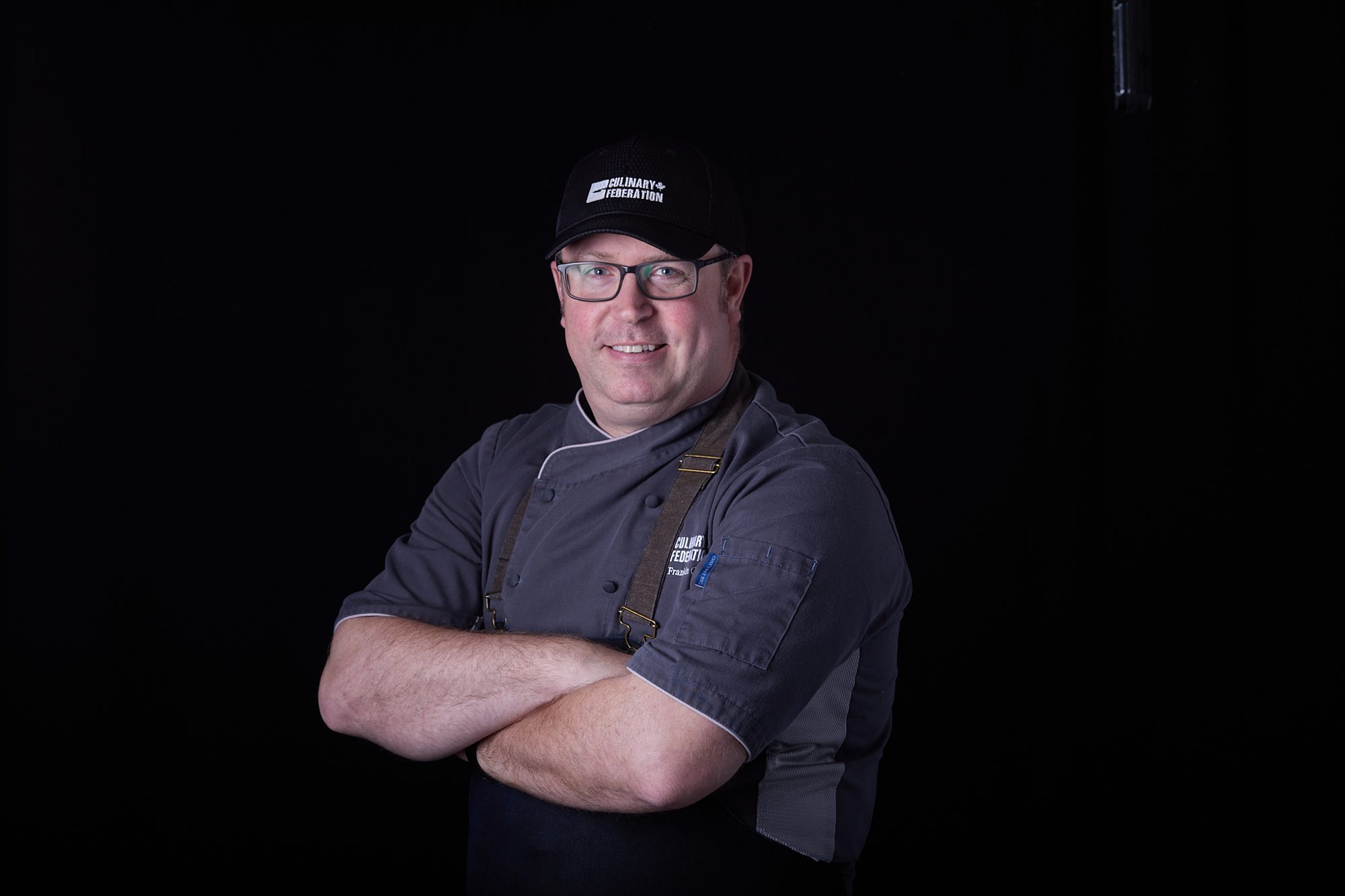 Chef in grey work shirt and cap, arms crossed, on black