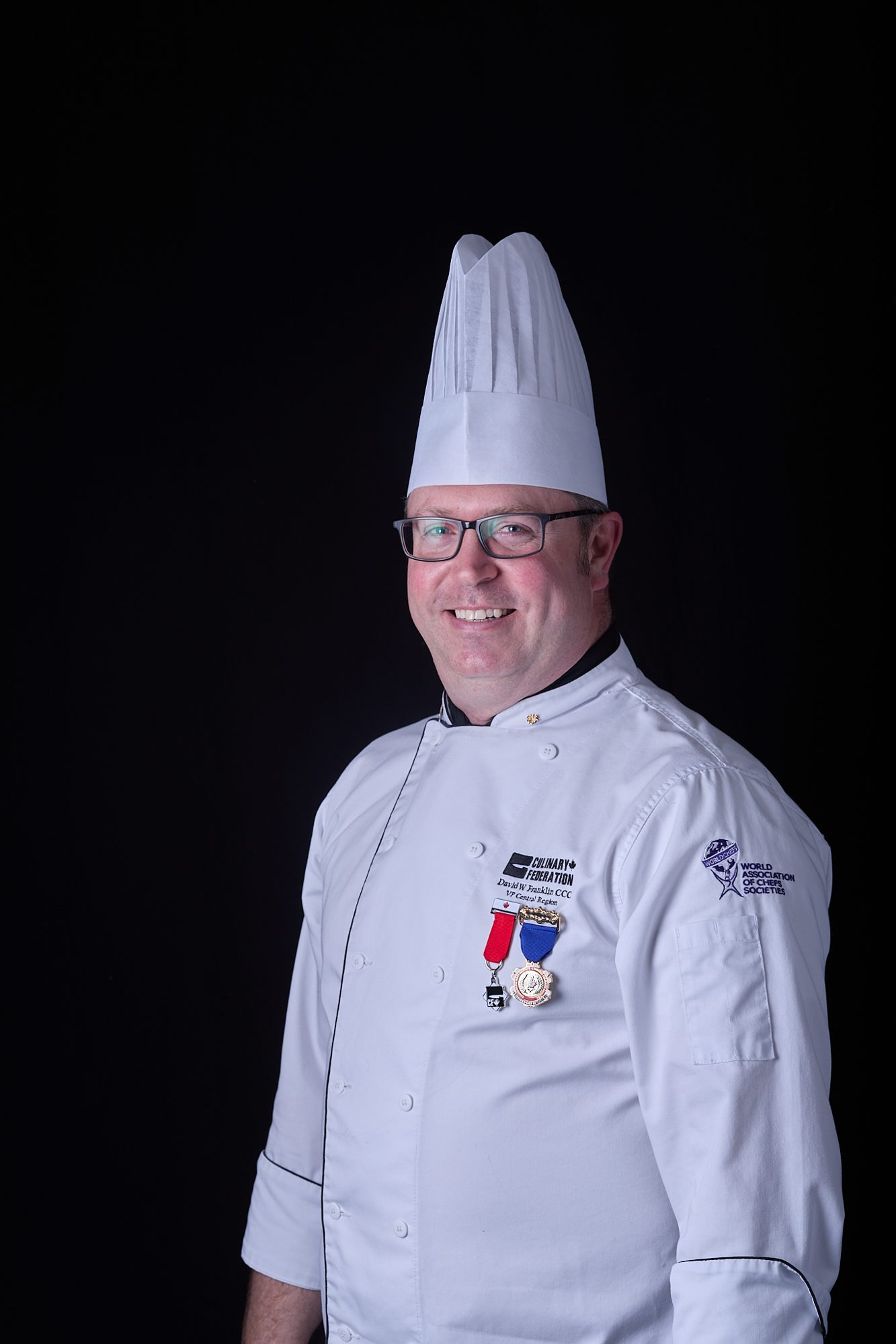Chef in formal whites with toque and medal, on black